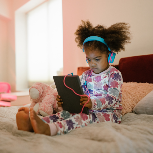 A child in her bedroom on an ipad, wearing headphones and looking comfy and cosy